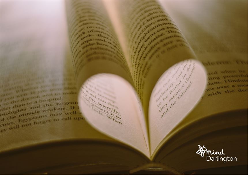 Photograph showing pages of an open book folded inward to form a heart shape at the center. Warm lighting highlights text on yellowed pages, creating a cozy and romantic atmosphere.