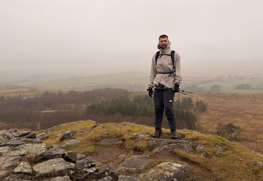 Photograph of a person standing on a rocky hilltop overlooking a foggy, expansive landscape with fields and sparse trees. The individual is dressed in outdoor hiking gear, including a beige jacket, black pants, gloves, and holding trekking poles, suggesting a hiking or trekking activity.
