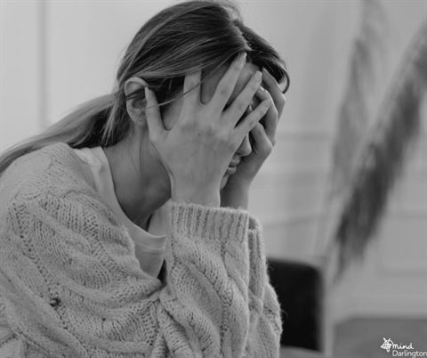 A person with long hair is sitting and holding their head with their hands, appearing distressed or overwhelmed. The image is in black and white, conveying a sense of emotional strain.